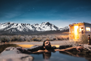 man in natural hot spring at night with snow capped mountains in the background